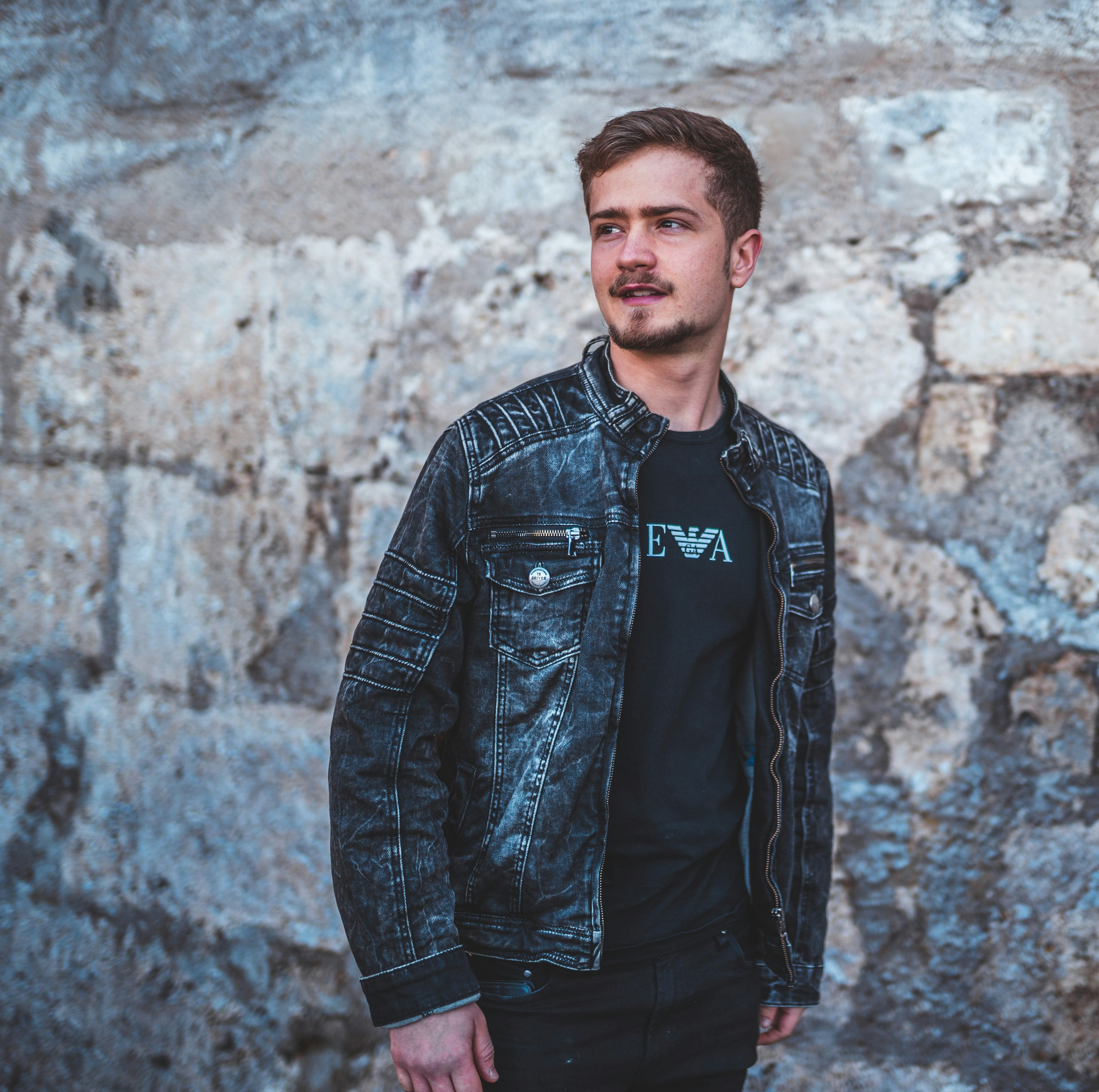 man in black leather jacket standing beside brown brick wall